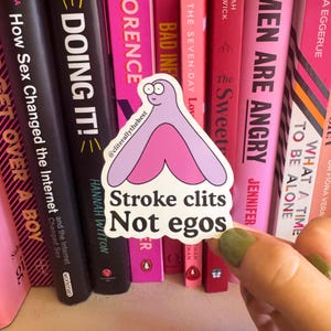 Può includere: Un adesivo con un'illustrazione a fumetti di una vulva in rosa e viola. L'adesivo ha il testo "Stroke clits Not egos". L'adesivo è tenuto davanti a uno sfondo di libri.