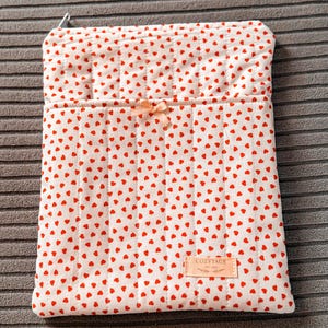 Puede incluir: Una funda acolchada blanca con un patrón de pequeños corazones rojos. La funda tiene un pequeño bolsillo y un lazo color melocotón. La etiqueta dice "COZYTAGE EST 2022".