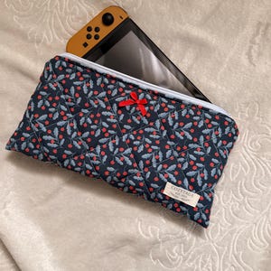 May include: A navy blue quilted case with a red berry and leaf pattern, holding a yellow and black game console. The case has a white zipper and a small red bow. The label says "COZYTAGE EST. 2023".