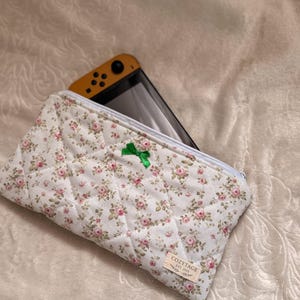 May include: A white quilted case with a floral pattern of pink roses and green leaves, containing a yellow and black handheld gaming device. The case features a green bow and a small tag reading "COZYTAGE EST. 2023".
