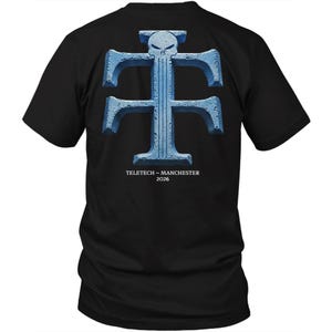 Könnte beinhalten: Schwarzes T-Shirt mit einem großen, hellblauen Aufdruck auf der Rückseite. Die Grafik ist ein stilisiertes "T" mit einem Totenkopf-Design oben. Unter der Grafik steht der Text "TELETECH - MANCHESTER 2026".