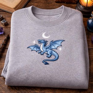 Peut inclure: Un sweat-shirt gris clair avec une broderie de dragon bleu, un croissant de lune et des étoiles. Le dragon est le motif central. Le sweat-shirt est plié sur une surface en bois.