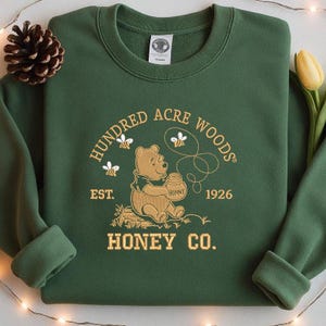 May include: Green sweatshirt with gold embroidery featuring Winnie the Pooh holding a honey pot. The text reads "Hundred Acre Woods Honey Co. Est. 1926". The design includes bees and a pinecone.