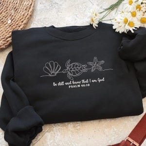 May include: Black sweatshirt featuring a white embroidered design of a seashell, sea turtle, and starfish. The text "Be still and know that I am God" and "PSALM 46:10" are also embroidered in white. The sweatshirt has long sleeves.