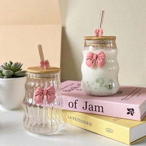 May include: Two clear glass tumblers with bamboo lids and pink bow details. One is empty, the other contains a white drink with green specks. Each lid has a gold-coloured straw and a small pink bow. The tumblers are placed on books.
