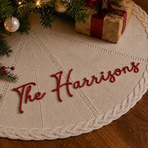 May include: A circular cream-coloured knitted Christmas tree skirt featuring a braided border and the red embroidered text The Harrisons. The fabric has a textured knit pattern with radiating lines extending from the centre to the outer edge.