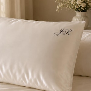 Puede incluir: Una funda de almohada rectangular de color marfil con un acabado suave y brillante. La tela presenta las letras del monograma JK bordadas en cursiva negra, posicionadas en la esquina superior derecha de la superficie de la funda de almohada.