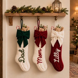 May include: Three cable-knit Christmas stockings hanging from a wooden shelf. The first is white with a green bow and the name Dad. The second is white with a red bow and the name Mom. The third is red with a white bow and the name Lilly.