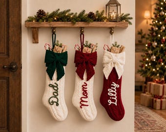 Könnte beinhalten: Drei gestrickte Weihnachtsstrümpfe hängen an einem Holzregal. Der erste ist weiß mit einer grünen Schleife und dem Namen Dad. Der zweite ist weiß mit einer roten Schleife und dem Namen Mom. Der dritte ist rot mit einer weißen Schleife und dem Namen Lilly.