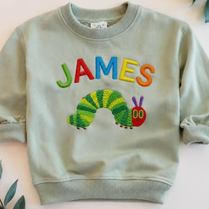 May include: A sage green long-sleeve sweatshirt featuring the name JAMES embroidered in multicolored letters above an embroidered green caterpillar with a red face and purple antennae. The garment has a ribbed crew neckline and ribbed cuffs.