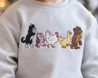 May include: A light gray crewneck sweatshirt featuring an embroidered line of animals across the chest. The design includes a black and white cat, a small purple owl, a white chicken, a yellow duck, and a brown bear walking in a single file line.