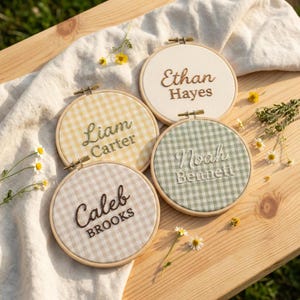 May include: Four embroidered name hoops with wooden frames. Each hoop features a different name: Ethan Hayes, Liam Carter, Noah Bennett, and Caleb Brooks. The fabric backgrounds have gingham patterns in various colors, including yellow, green, and beige.