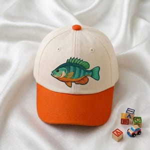 May include: A baseball cap with an off-white crown and a solid orange brim. The front panel features an embroidered graphic of a green and orange fish with dark stripes. The cap has a small orange button at the top center of the crown.