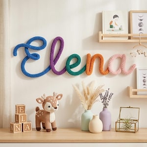 May include: A colorful, knitted "Elena" name sign in blue, purple, green, orange, and pink is mounted on a white wall. A crocheted deer, wooden blocks spelling "ELNA", and decorative vases are on a wooden shelf.