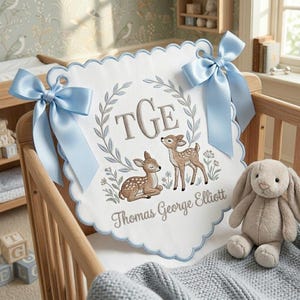 May include: A white scalloped fabric banner featuring two embroidered deer, a laurel wreath, the monogram TGE, and the name Thomas George Elliott. The banner is decorated with two large, light blue satin bows attached to the top corners.