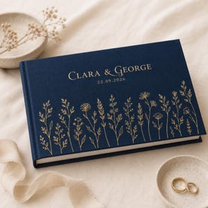 May include: A navy blue hardcover guest book featuring gold foil botanical illustrations along the bottom edge. The cover is personalized with the text Clara & George and the date 22.09.2026 printed in a gold serif font centered at the top.