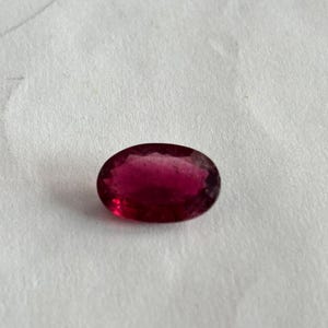 May include: An oval-cut, deep red gemstone. The polished stone has a smooth surface and is set against a white background. The gem's colour ranges from a dark, saturated red to a lighter, translucent hue.