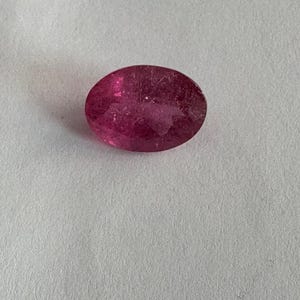 Peut inclure: Une pierre précieuse rose, de forme ovale et facettée. La gemme a une couleur profonde et saturée et semble translucide. La pierre est posée sur un fond blanc. Cette pierre précieuse pourrait être utilisée en joaillerie.