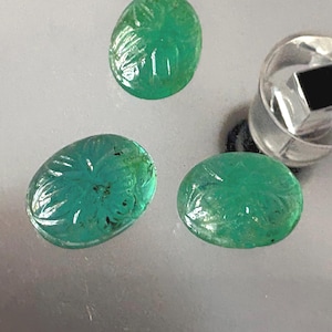 May include: Three oval-shaped, carved emerald gemstones. The gemstones are a vibrant green color and feature intricate floral or leaf-like carvings on their surfaces. A silver-colored object is in the background.