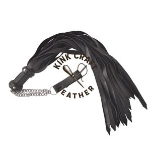 Pode incluir: Flogger de couro preto com várias tiras longas e finas. A pega é cilíndrica com uma corrente e um anel prateados. O logótipo "KINK CRAFT LEATHER" é visível.