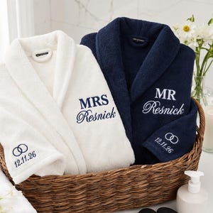May include: A white bathrobe embroidered with navy blue text reading MRS Resnick and a navy blue bathrobe embroidered with white text reading MR Resnick, both featuring a wedding ring icon and the date 12.11.26 on the sleeve cuffs.