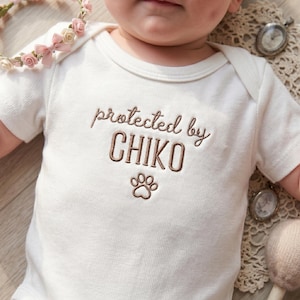 May include: A white baby onesie with short sleeves, featuring the embroidered text "protected by CHIKO" in brown thread, along with a paw print. A floral crown with pink roses is visible in the upper left corner.