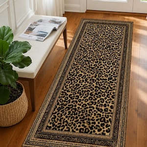May include: A rectangular runner rug featuring a prominent leopard print pattern in shades of tan, brown, and black. The rug is framed by a dark, intricate border with a repeating geometric design, creating a distinct contrast against the central animal print.