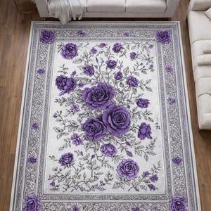 May include: Rectangular area rug with a floral design. The rug features a white background with a pattern of purple roses and green leaves. A gray border with floral detailing frames the design. The rug is placed on a wooden floor.