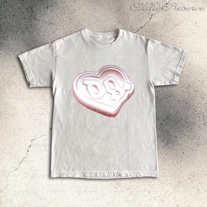 May include: A white t-shirt featuring a heart-shaped graphic with a 3D effect. The graphic has a white center with a pink outline and the letters "DQ" inside. The shirt is made of a soft material and has short sleeves.