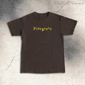 Peut inclure: Un t-shirt marron avec le mot "Pinegrove" imprimé en jaune sur la poitrine. Le t-shirt a des manches courtes et un col rond. Le texte est dans une police simple sans empattement.