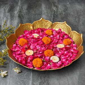 Peut inclure: Un bol en métal doré en forme de fleur de lotus, rempli de pétales de rose rose vif, de plusieurs fleurs de souci orange et de trois petites bougies chauffe-plat blanches allumées, formant un centre de table décoratif.