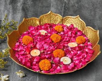 Puede incluir: Un cuenco de metal martillado en tono dorado con forma de flor de loto, lleno de pétalos de rosa de color rosa vibrante, flores de caléndula de color naranja brillante y tres pequeñas velas de té blancas redondas con mechas encendidas.