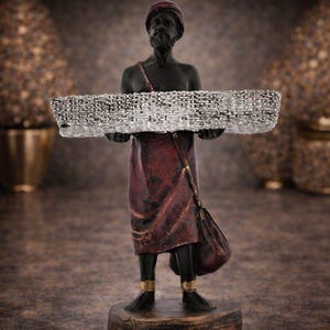 May include: A dark-skinned figurine of a man in a turban and maroon robe, holding a silver-coloured basket. The statue has gold accents on the ankles and a matching shoulder bag. The base is a dark brown colour.