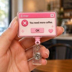 May include: A rectangular pink and white badge reel featuring a computer error message graphic. The text reads Error, You need more coffee, and OK. The design includes a pink circle with an X, a pink heart, and small four-pointed star icons.