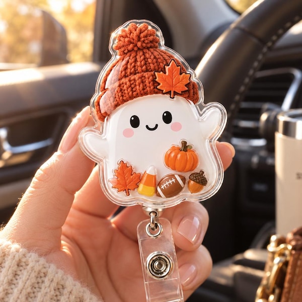 May include: A clear acrylic badge reel featuring a white ghost character wearing a textured orange knit beanie with a maple leaf accent. The ghost is decorated with small autumn-themed charms including a pumpkin, candy corn, football, and acorn.