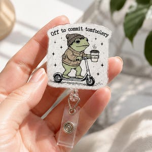 May include: A badge reel featuring a square acrylic topper with a speckled white background. The illustration shows a green frog wearing a brown sweater and grey beanie, riding a scooter while holding a coffee cup labeled Caffeine & Chaos. The text above reads Off to commit tomfoolery.