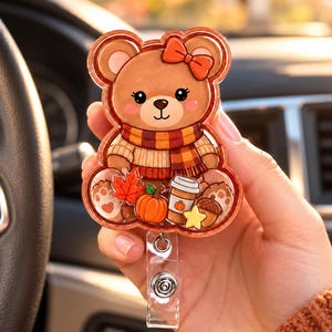 May include: A bear-shaped badge reel featuring a brown bear wearing a red bow and a striped scarf. The design includes a red maple leaf, an orange pumpkin, a white coffee cup with a pumpkin icon, a yellow star, and a brown acorn at the base.