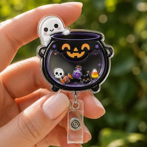 May include: A clear acrylic badge reel featuring a black cauldron with an orange jack-o'-lantern face. Inside the cauldron are small floating charms including a white skull, a purple potion bottle, a black cat, a candy corn, and an orange candy.