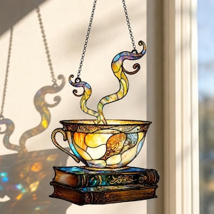May include: A stained glass hanging depicting a teacup with steam rising from it, resting on two books. The teacup is a mix of yellow, orange, and blue glass. The steam is a swirling design of yellow, blue, and purple. The books are brown with blue and green accents.