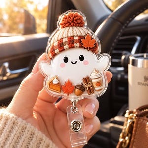 May include: A clear acrylic badge reel featuring a white ghost character wearing a knit beanie with a pom-pom and a maple leaf. The ghost is decorated with miniature autumn-themed charms including a slice of pumpkin pie, a leaf, an acorn, cinnamon sticks, and a coffee cup.