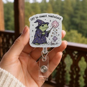 May include: A badge reel featuring a light gray rectangular base with a green frog wearing a purple wizard hat and robe decorated with yellow stars. The frog holds a wand emitting pink smoke and stars. The text reads Off to commit tomfoolery.
