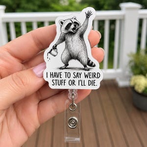 May include: A badge reel featuring a black and white illustration of a raccoon waving one paw while holding a small mug in the other. Below the raccoon, the text reads: I HAVE TO SAY WEIRD STUFF OR I'LL DIE. The badge reel has a clear plastic strap.