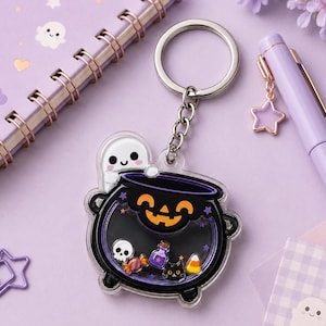 May include: A clear acrylic shaker keychain featuring a black cauldron with an orange jack-o'-lantern face. Inside the cauldron are small floating charms including a white skull, a purple potion bottle, a black cat, and candy corn. A small white ghost sits on the rim.