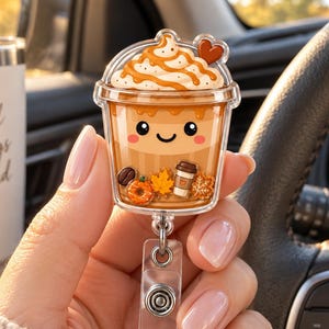 May include: A clear acrylic badge reel featuring a cartoon iced coffee cup with whipped cream, a small heart, and autumn-themed charms including a pumpkin, a maple leaf, and a tiny coffee cup, all set against a light brown beverage background.