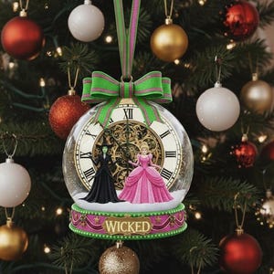 May include: A clear glass Christmas ornament with a clock face design, featuring figures of two characters from the musical "Wicked." The ornament has a green and pink ribbon and bow. The word "WICKED" is displayed on the base.