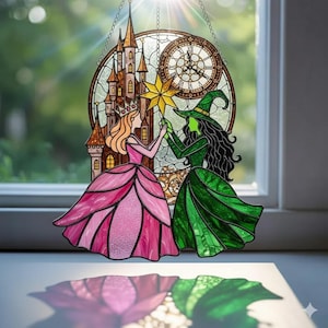 Può includere: Suncatcher in vetro colorato con una principessa e una strega che tengono una stella. Il design include un castello, un orologio e una combinazione di colori verde e rosa. Il suncatcher è appeso davanti a una finestra.