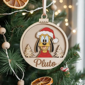 May include: Wooden Christmas bauble featuring Pluto in a Santa hat and red sweater. The circular design includes Pluto's image and the name "Pluto" engraved below. Two small Christmas trees are also included. The bauble hangs from a beige ribbon, displayed on a Christmas tree.