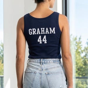 Könnte beinhalten: Marineblaues Tanktop mit dem Namen "GRAHAM" und der Nummer "44" in weißer Schrift. Das Tanktop wird mit hellblauen Jeans getragen. Die Person ist von hinten zu sehen.