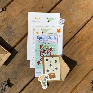 Op de afbeelding: Een assortiment briefpapier, waaronder een gastenbon, een kaart met een blauwe bessenontwerp en een sticker met een bloemenillustratie, gerangschikt op een houten oppervlak. De items hebben een vintage esthetiek met handgeschreven lettertypen en stermotieven.