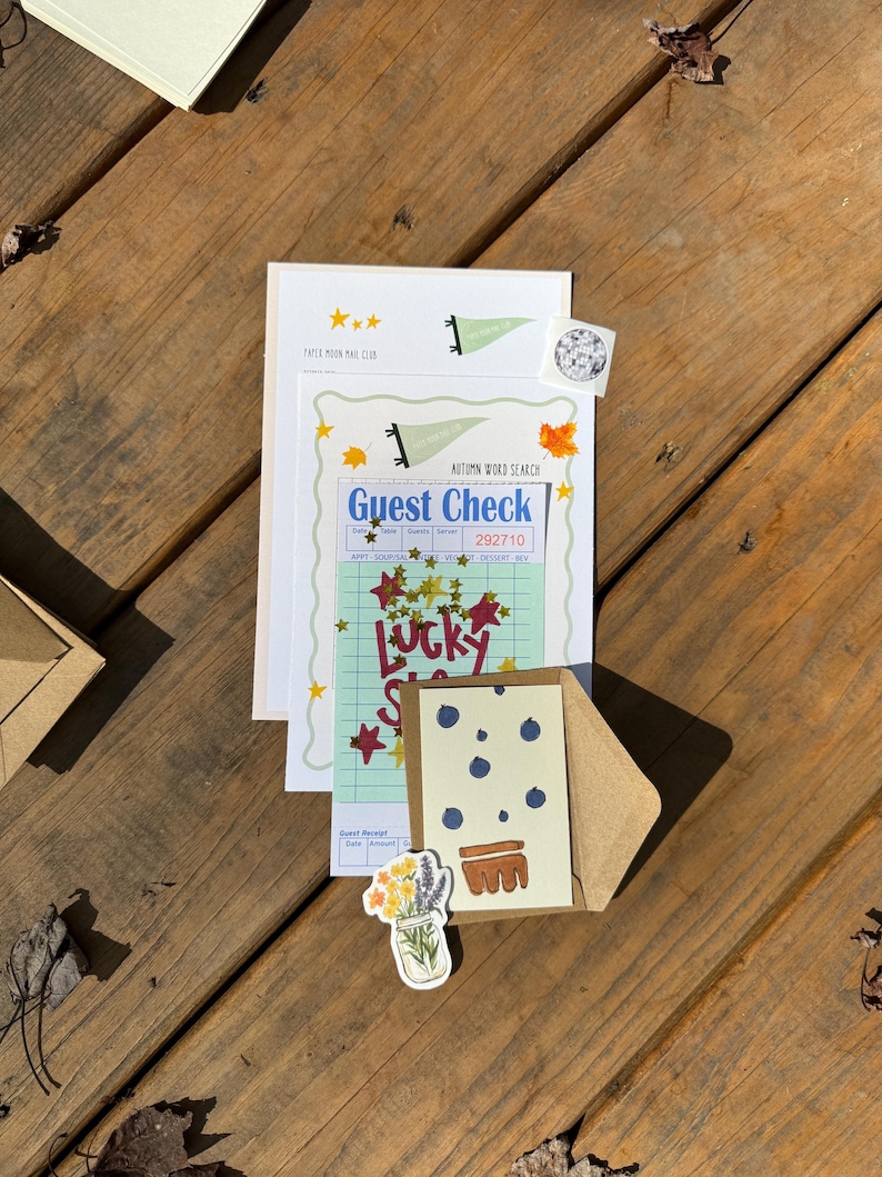 Op de afbeelding: Een assortiment briefpapier, waaronder een gastenbon, een kaart met een blauwe bessenontwerp en een sticker met een bloemenillustratie, gerangschikt op een houten oppervlak. De items hebben een vintage esthetiek met handgeschreven lettertypen en stermotieven.